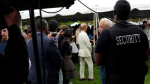 This photo shows a security guard standing outdoors near a crowd of eventgoers.